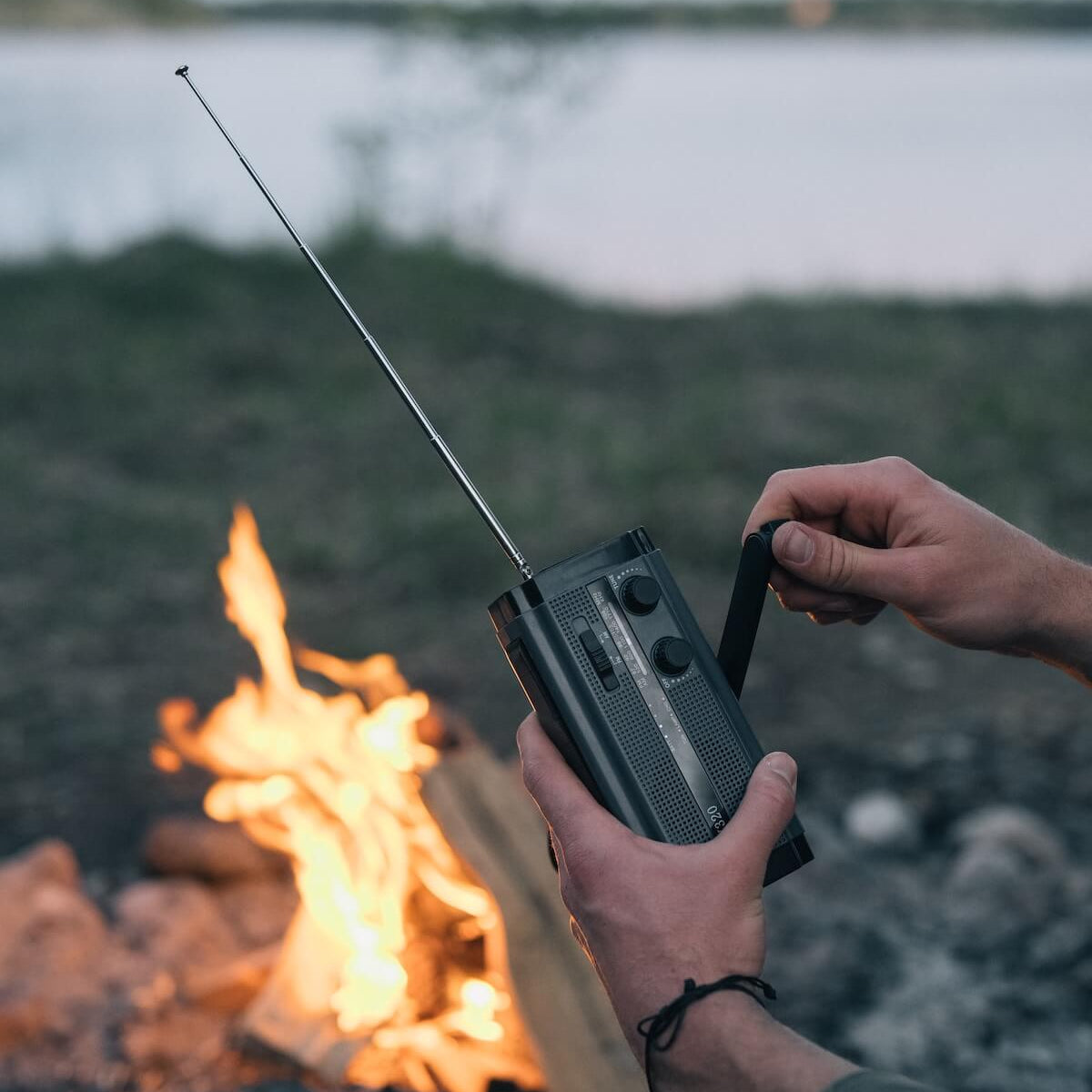 En person skruer på en sort håndsvingradio med en udfoldet antenne, med et lejrbål og en sø i baggrunden.