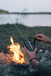 En person skruer på en sort håndsvingradio med en udfoldet antenne, med et lejrbål og en sø i baggrunden.