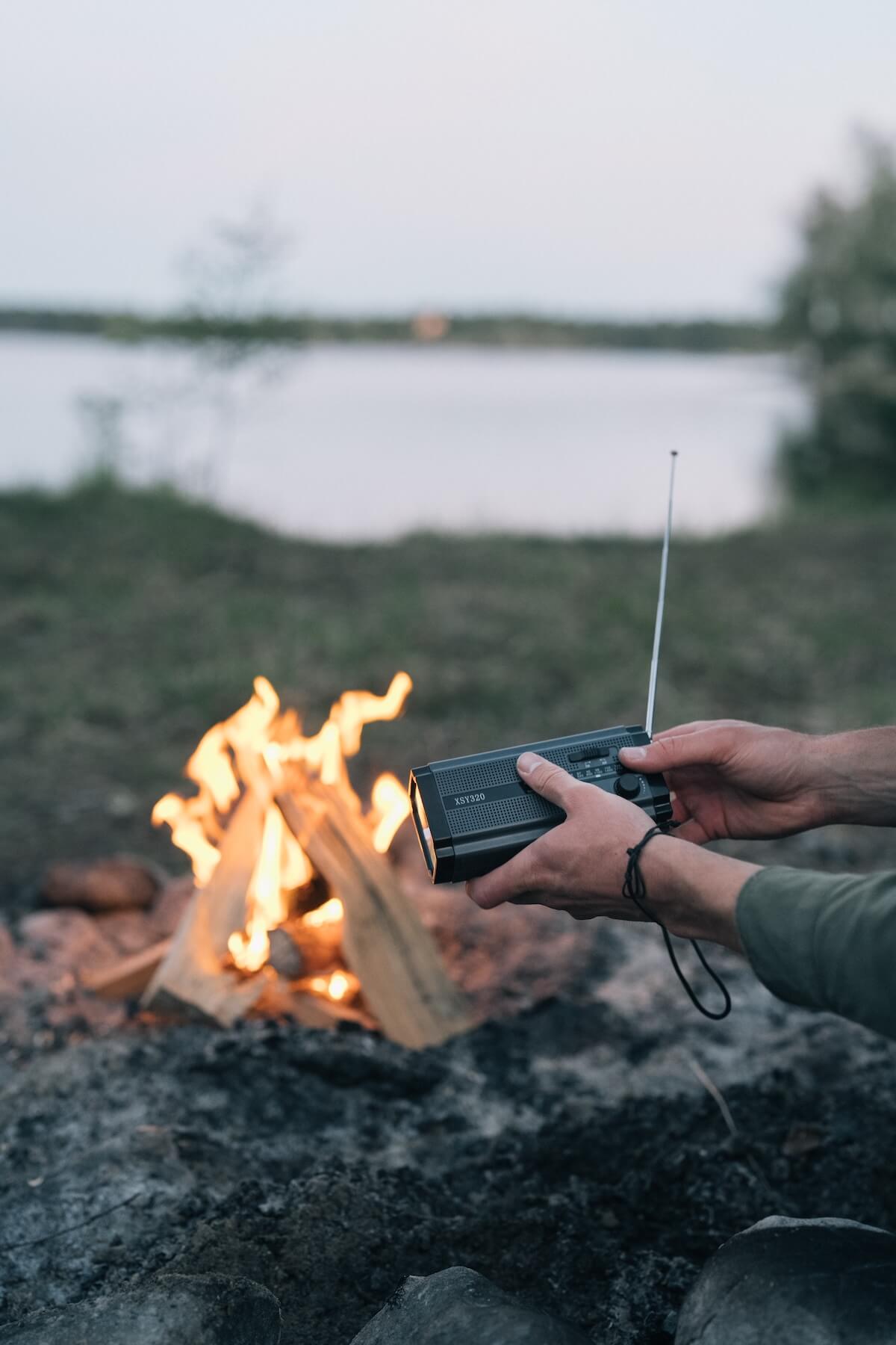 En person holder en sort håndsvingradio med en udfoldet antenne ved siden af et knitrende lejrbål med en sø i baggrunden.