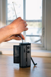 En hånd justerer antennen på en sort håndsvingradio med solpanel, der er placeret på et træbord foran et vindue.