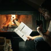 En person läser en bok vid en sprakande eldstad, upplyst av en ficklampa.