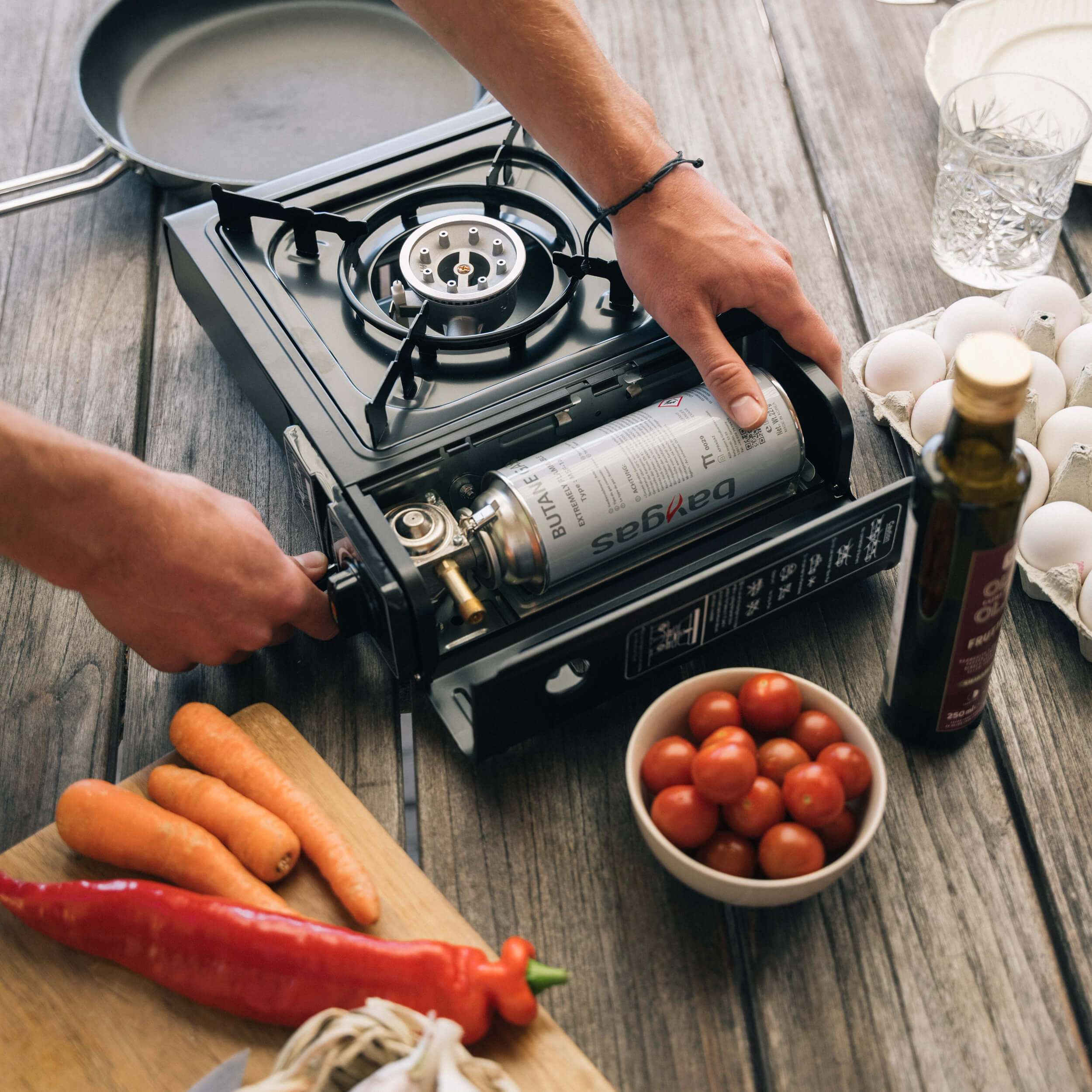 En person sätter in en gaspatron i ett litet gasolkök på ett träbord med grönsaker och ägg.