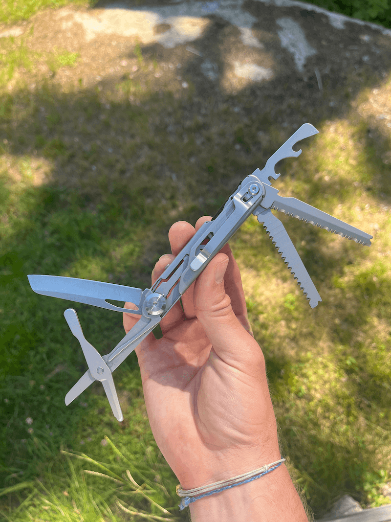 En hånd holder et udfoldet multiværktøj med kniv, sav og proptrækker op mod en græsplæne i baggrunden.