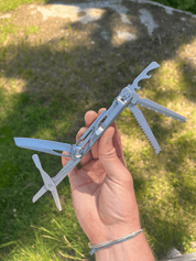 En hånd holder et udfoldet multiværktøj med kniv, sav og proptrækker op mod en græsplæne i baggrunden.