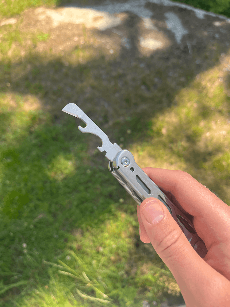 En hånd holder et multiværktøj med en udfoldet oplukker mod en frodig grøn græsplæne i solskin.