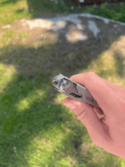 Den ene hånd holder et foldet multiværktøj mod en frodig grøn græsplæne med solpletter.