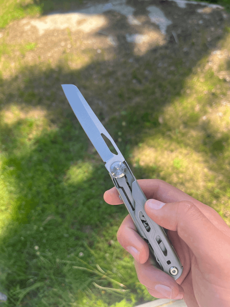 En hånd holder et multiværktøj med en udfoldet kniv på en solbeskinnet græsplæne med skygger.