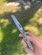 En hånd holder et multiværktøj med en udfoldet kniv på en solbeskinnet græsplæne med skygger.