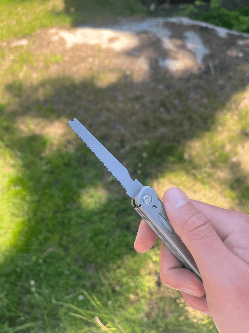 En hånd holder et multiværktøj med en udfoldet sav på en solrig græsplæne med skygger.