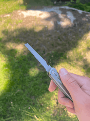 En hånd holder et multiværktøj med en udfoldet sav på en solrig græsplæne med skygger.