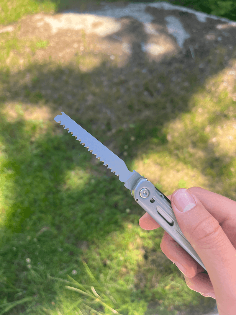 En hånd holder et multiværktøj med en udfoldet sav på en græsklædt baggrund.