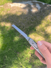 En hånd holder et multiværktøj med en udfoldet sav på en græsklædt baggrund.