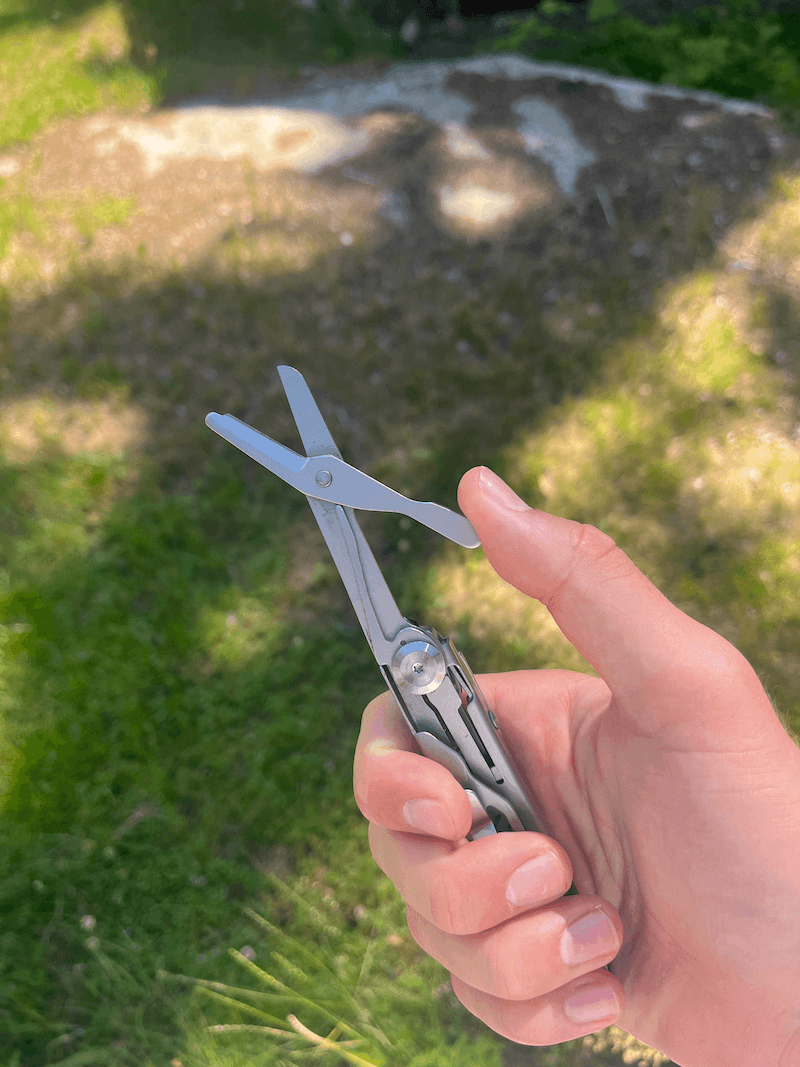 En hånd holder et multiværktøj med en udfoldet saks mod en grøn græsplæne i sollys.