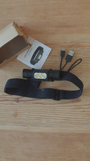 En svart pannlampa med USB-laddningskablar och en bruksanvisning på ett träbord.