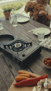 Ett gasolkök på ett bord utomhus, med ägg, grönsaker, porslin och en hund i bakgrunden.