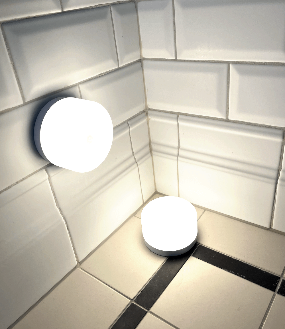 Två runda, batteridrivna lampor som lyser i ett hörn med vita kakelplattor.