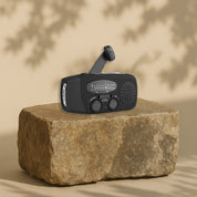 En svart vevradio med solpanel och ficklampa, placerad på en stenblock med skuggor i bakgrunden.