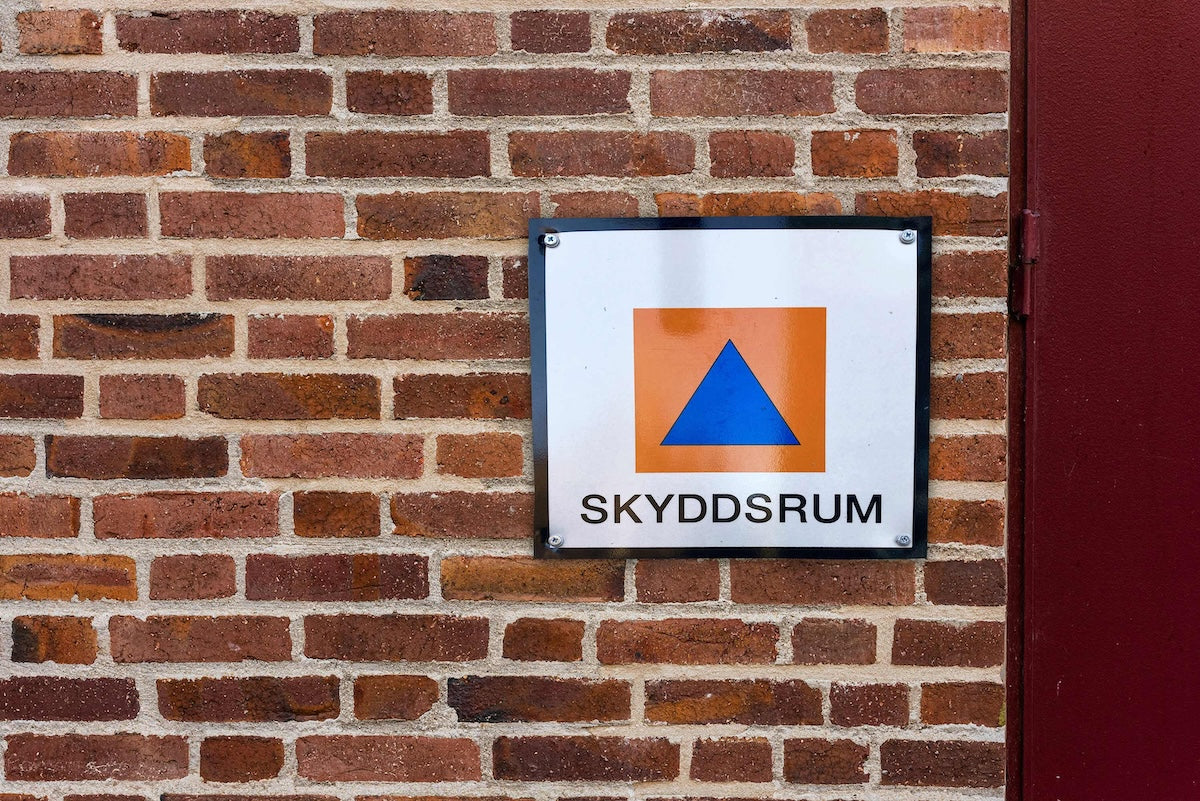 Skyddsrumsskylt med orange fyrkant och blå triangel på en tegelvägg