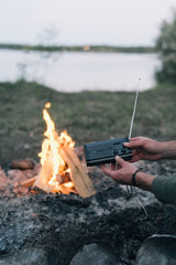 En person håller en svart vevradio med utfälld antenn bredvid en sprakande lägereld, med en sjö i bakgrunden.