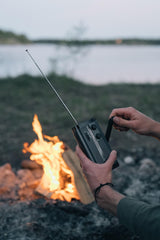 Henkilö pyörittää mustaa kampiradiota, jonka antenni on auki, ja taustalla on nuotio ja järvi.