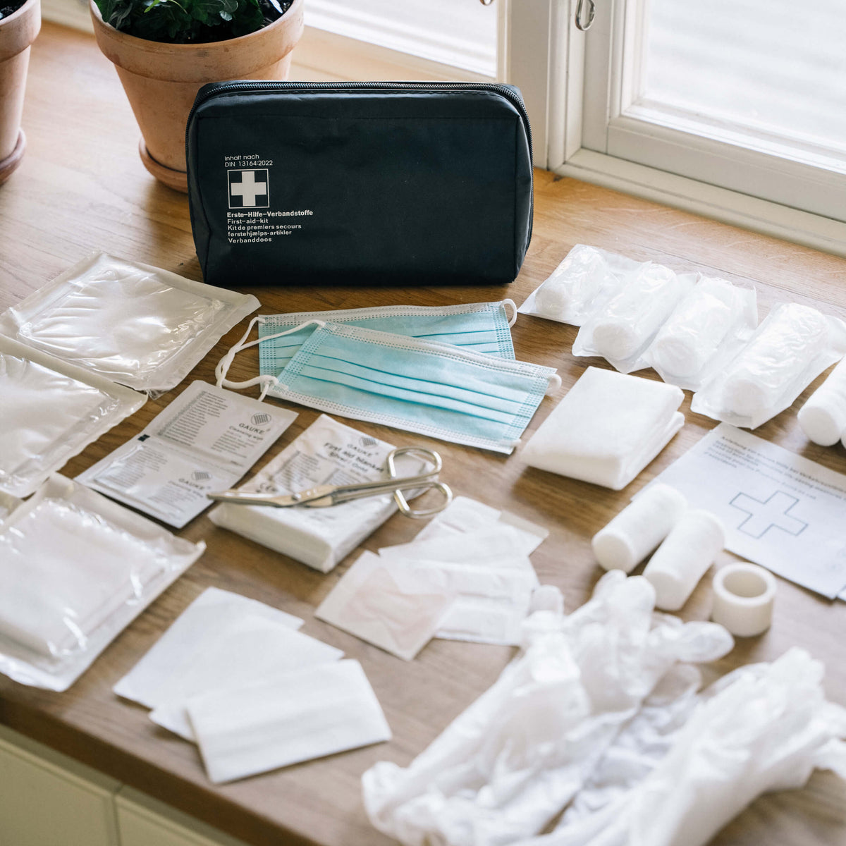 Ett svart första hjälpen-kit har öppnats och innehållet, inklusive gasbindor, sax, masker och handskar, ligger utspritt på ett träbord.