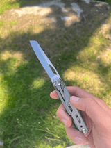 En hand håller ett multiverktyg med en utfälld kniv, mot en solig gräsmatta med skuggor.