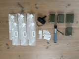 Krislådans innehåll uppställt på ett trägolv: hopfällbara vattenbehållare, en vevradio, vakuumförpackade nödponchos, vattenreningstabletter från Katadyn och ett portabelt vattenfilter