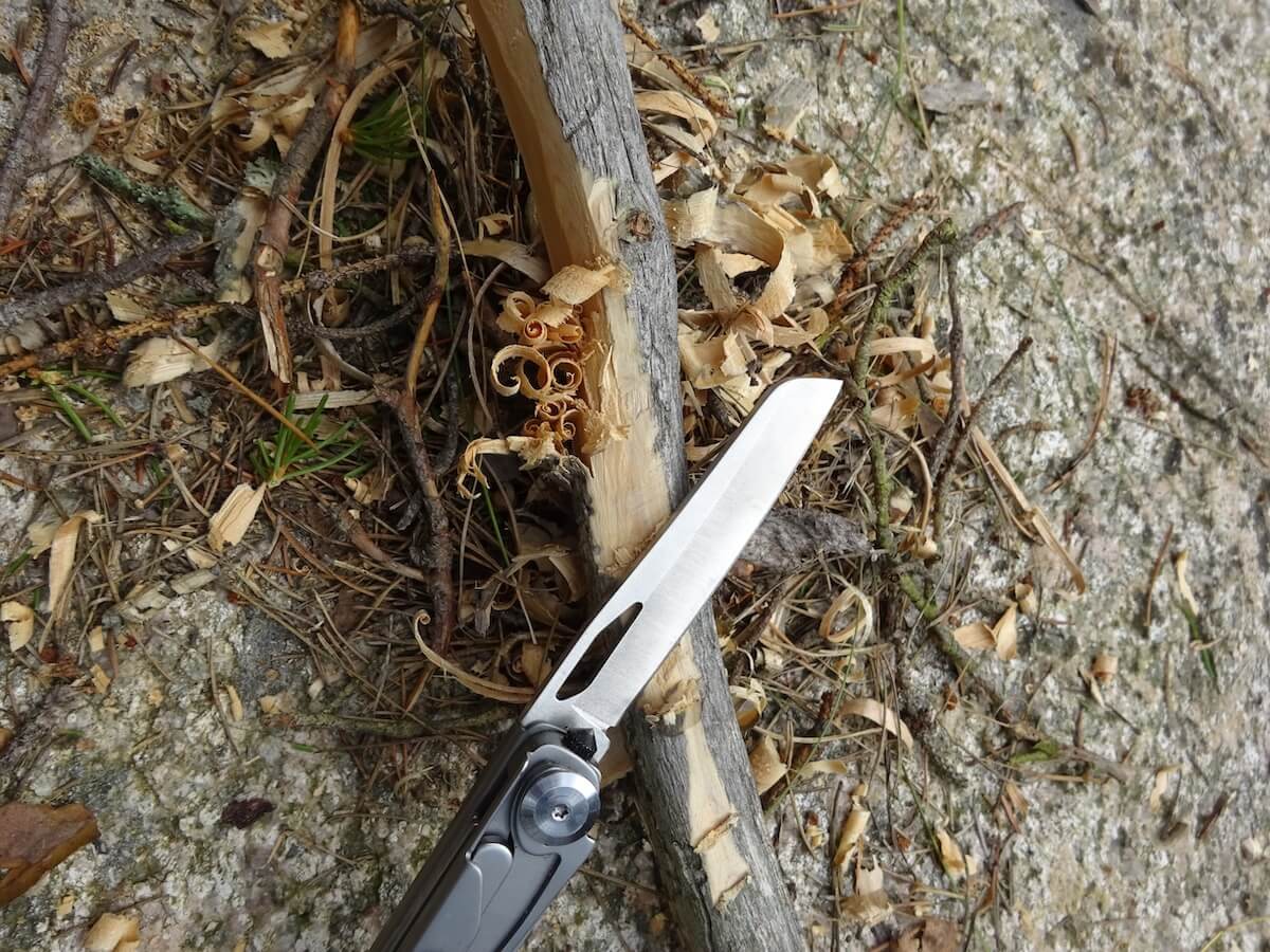 En multiverktygskniv skär i en träpinne, med träflisor utspridda på marken.